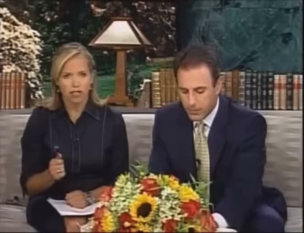 The video opens on two news anchors seated at a broadcast desk before transitioning to a wide camera shot of the New York City skyline featuring the Empire State Building in the midground and the World Trade Center towers in the background. Dense dark smoke is pouring out from the upper levels of the North Tower. A female news anchor announces that a plane has reportedly crashed into the World Trade Center and introduces a phone witness.